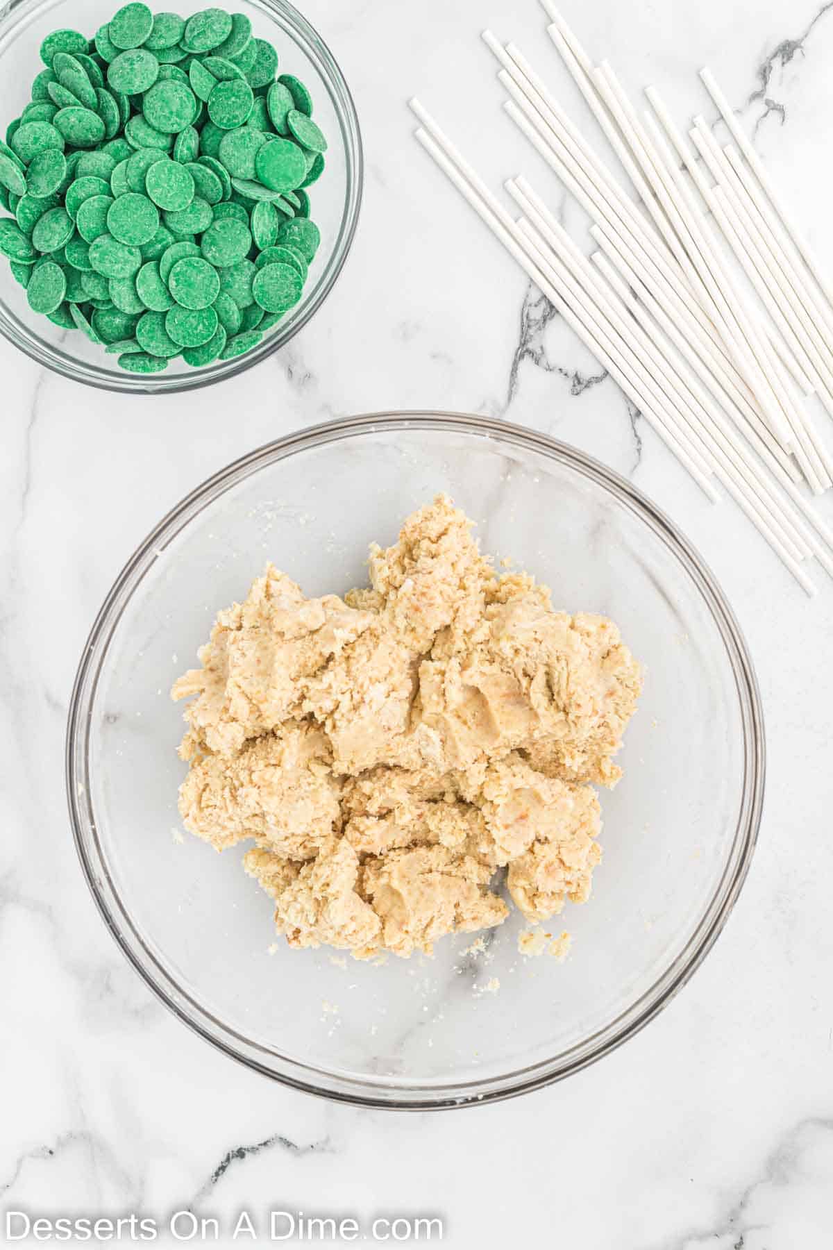 Frosting mixed with cake crumbles in a bowl and a small bowl of green candy melts and lollipop sticks
