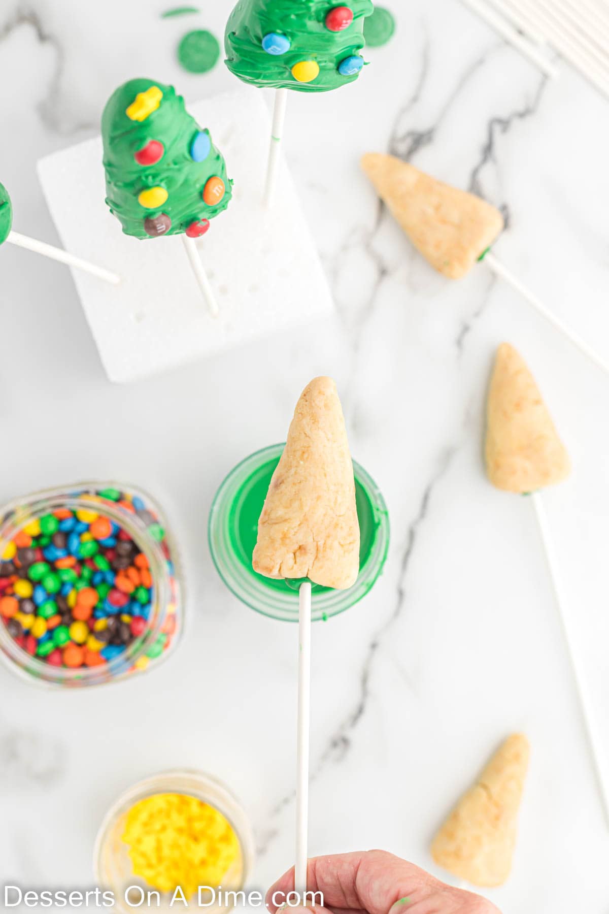 Corn shaped cake mixture on a lollipop stick with melted candy melts in a small bowl with a jar of M&M's
