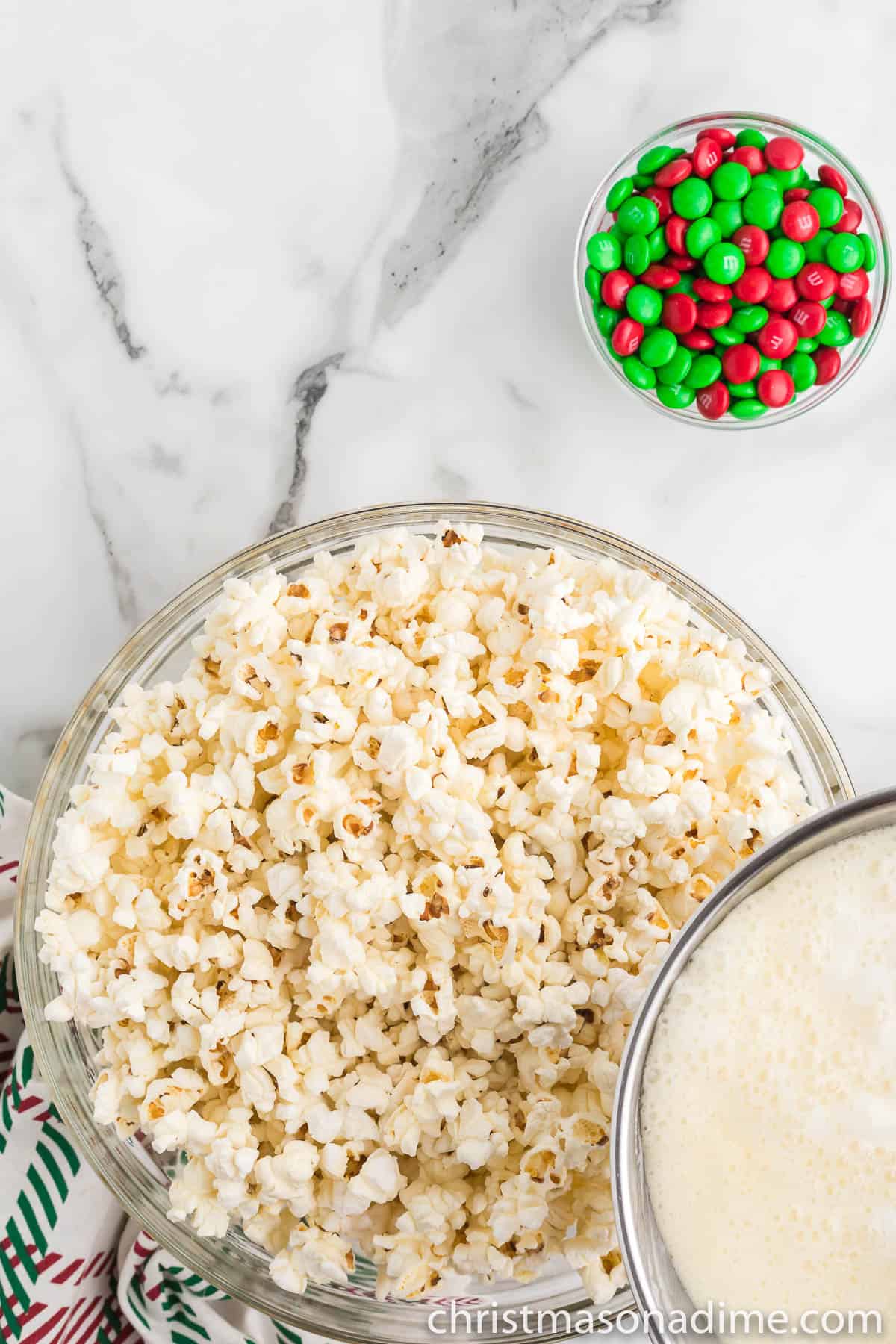 Pouring the melted marshmallow mixture over the popcorn in a bowl with a small bowl of M&Ms on the side