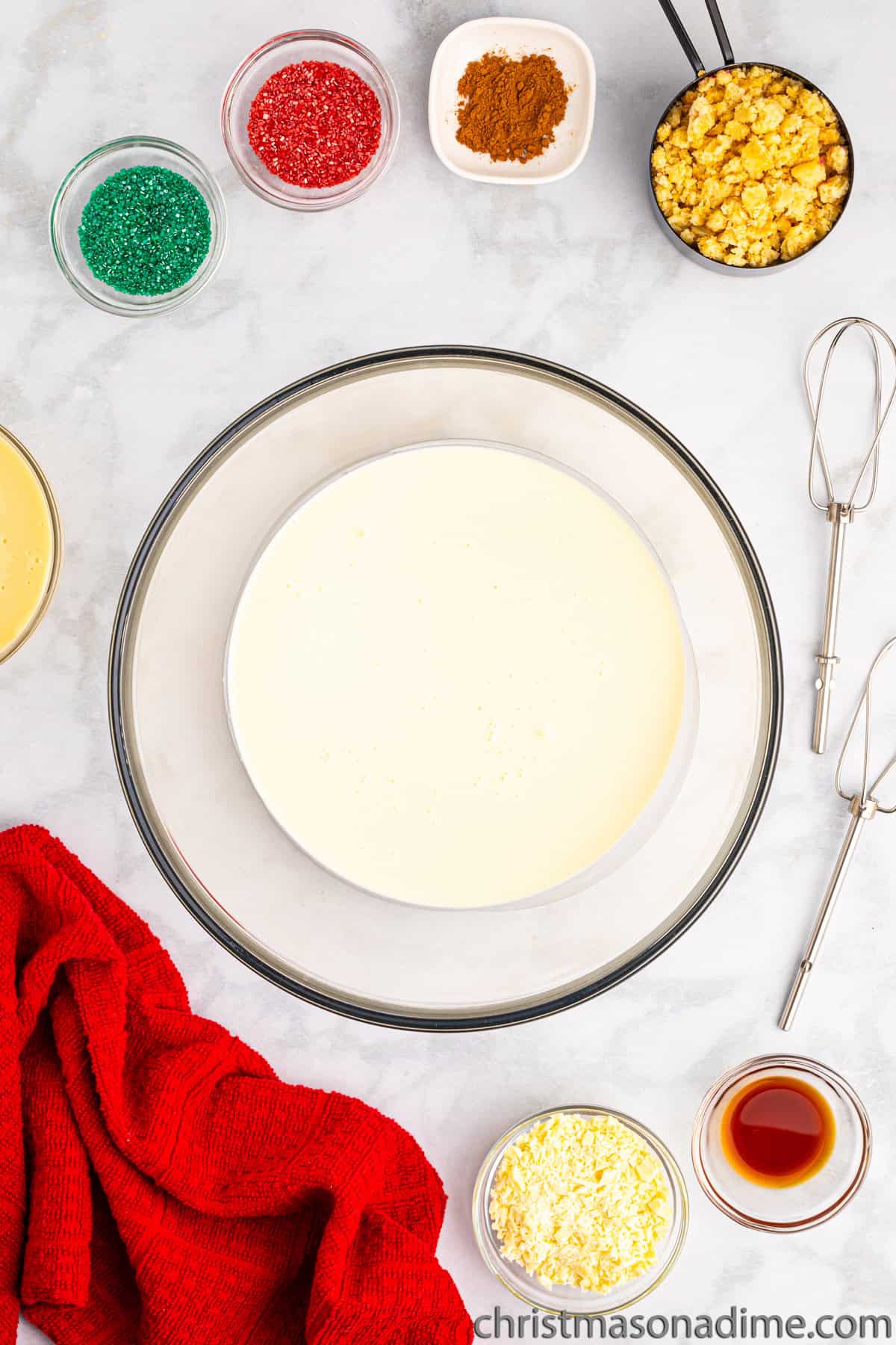 Heavy Cream in a bowl with small bowls of red and green sprinkles, vanilla, cinnamon and crushed cookies